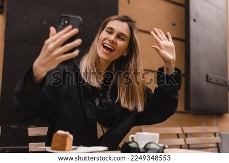 Happy woman using smartphone at cafe. Smiling blonde woman using smart phone make selfie and make hello gesture waving hand, spends her leisure time in cafe, video call. Blonde girl wear black shirt.