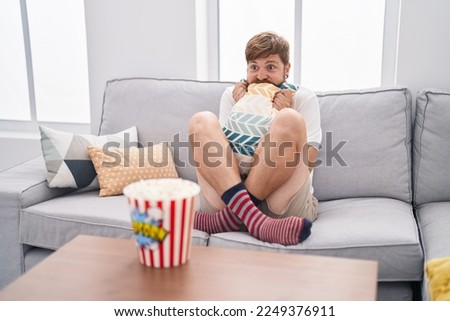 Young redhead man watching movie with scare expression at home