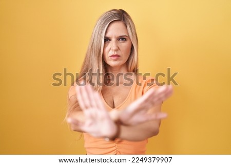 Young woman standing over yellow background rejection expression crossing arms and palms doing negative sign, angry face 