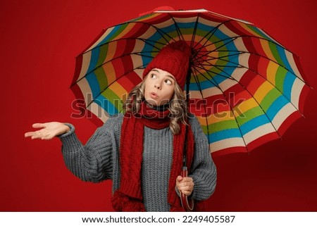 Young woman wear grey sweater scarf hat hold opened colorful umbrella catch raindrops isolated on plain red background studio portrait. Healthy lifestyle ill sick disease treatment cold season concept