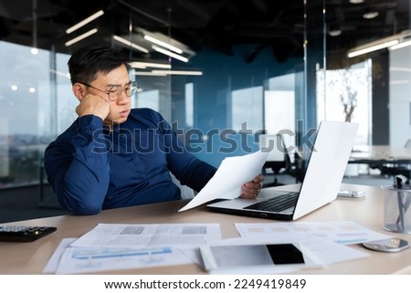 Tired young Asian man, accountant, businessman, freelancer. He sits in the office at a table with a laptop, looks through documents, holds his head in his hands, sighs a hard and boring day. Royalty-Free Stock Photo #2249419849