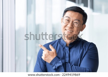 Close-up photo. Portrait of a young male Asian teacher. It stands in the office, on the campus near the window. He looks at the camera, smiles, makes a success gesture with his finger.