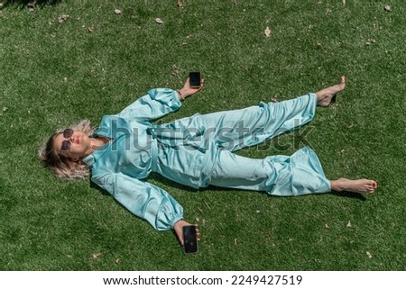 A woman with light curly hair wearing glasses and a blue jumpsuit is lying on the summer fresh green grass with a phone in her hands.
