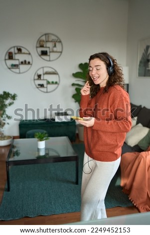 Happy pretty young woman wearing headphones dancing at home. Cheerful girl listening music on mobile phone, singing song feeling relaxed standing in modern living room interior. Vertical.