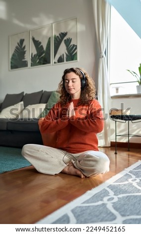 Young healthy mindful woman wearing headphones listening calming music audio podcast sitting on floor at home doing yoga meditation relaxing exercise for mental balance feeling peace of mind. Vertical Royalty-Free Stock Photo #2249452165