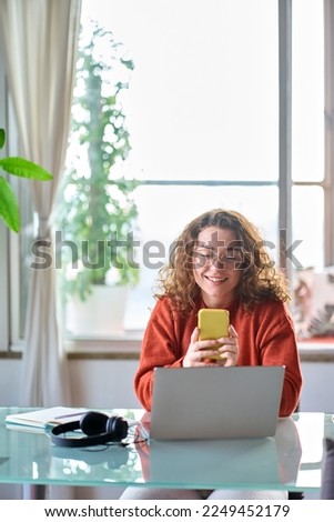 Young happy pretty woman sitting at table holding smartphone using applications on cellphone modern technology, looking at mobile phone while working or learning at home. Vertical