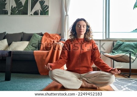 Young pretty healthy mindful woman wearing headphones listening calming music audio podcast sitting on floor at home doing yoga meditation relaxing exercise for mental balance in apartment interior. Royalty-Free Stock Photo #2249452183
