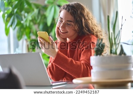Young happy smiling pretty woman holding smartphone using cellphone modern technology, looking at mobile, checking cell phone apps, watching social media sitting at table at home.