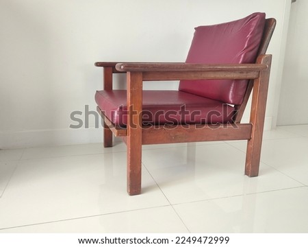 Old wooden chair. There is a red seat and backrest, not very high but very strong, made of brown solid wood and tanned hides. Set on a tiled floor and a white background wall. Used for long time.
