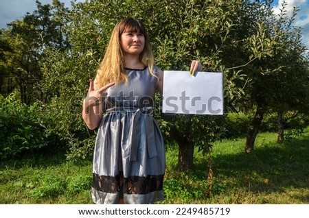 Front view of smiling young stylish brunette girl pointing at white paper sheet in her hand posing on nature background. Concept of advertising.
