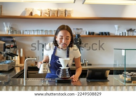 Happy smiling asian girl smelling coffee while brewing filter, standing behind counter in cafe.