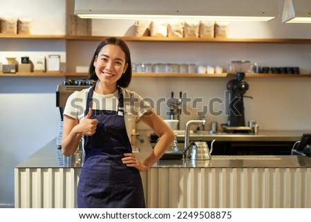 Smiling girl student working part-time in cafe, barista shows thumbs up, wears apron, stands near coffee shop counter. Royalty-Free Stock Photo #2249508875