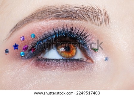 Closeup macro shot of human female brown color eye. Woman with natural evening vogue face beauty makeup. Girl with perfect skin, with blue eye shadow make up and rhinestones. Royalty-Free Stock Photo #2249517403