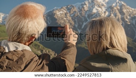 Senior caucasian couple having a rest on top of a mountain, taking a picture on smartphone, travelling together after retirement - pension, recreational pursuit, tourism concept 