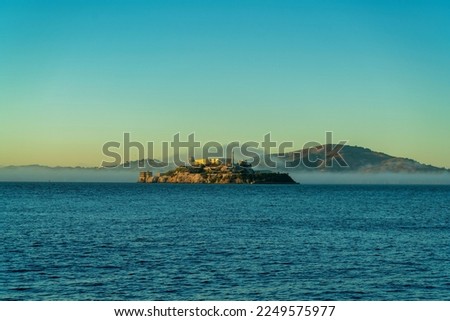 Distant alcatraz insland on the ocean with mist and fog and background mountains in golden sunset or sunrise. Near the beach with clear blue sky with buildings on prison in san francisco california.