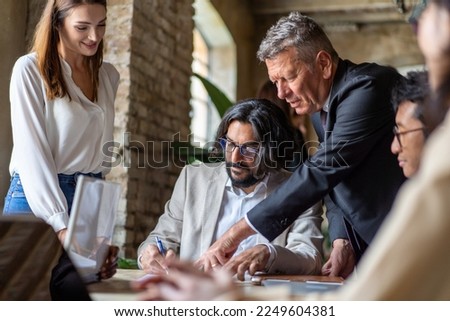 Arab man signs a contract guided by an older man in a suit while a diverse group of people look on. Multicultural team works together to finalize a business deal, diversity and collaboration concept