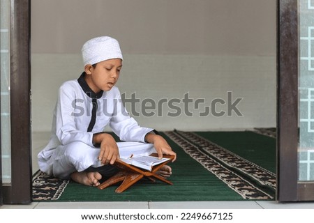 Muslim kid using white rob and skullcap is sitting and reciting Al Quran at the mosque.  Royalty-Free Stock Photo #2249667125