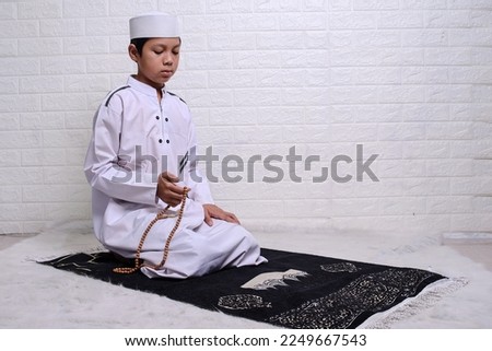 Muslim kid wearing white rob and skullcap is sitting and holding prayer beads or tasbih at prayer mat  Royalty-Free Stock Photo #2249667543