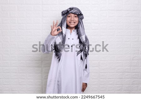 Smiling muslim kid wearing white rob and turban showing ok sign on white background. 