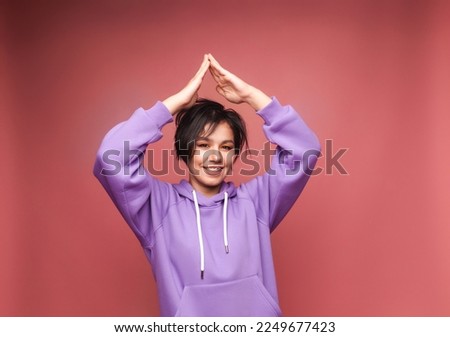 A happy young girl with short black hair makes a gesture of the roof of the house with her hands over her head. The concept of mortgage insurance. isolated on a pink background