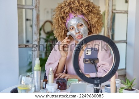 Photo of young curly female model keeps lips rounded applies facial silver mask uses brush for making massage tries different cosmetic products records video blog tells how to care about skin
