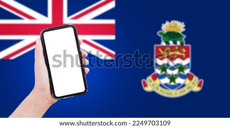 Close-up of male hand holding smartphone with blank on screen, on background of blurred flag of Cayman Islands.