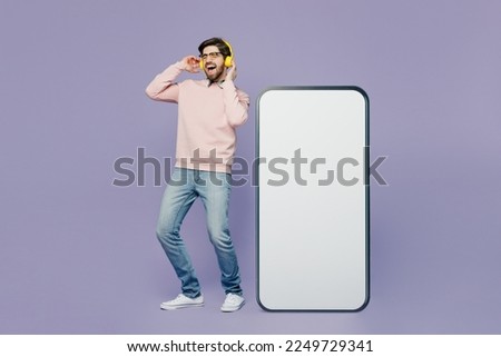 Full body young IT man he wear casual clothes pink sweater glasses big huge blank screen mobile cell phone smartphone with area listen to music in headphones isolated on plain pastel purple background