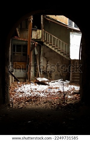 Vertical photo. View from a dark unlit arch to the courtyard of the house. Old broken wooden staircase and door. The concept of desolation, abandonment, loneliness, depression, decline of culture.