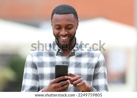 Front view portrait of a happy black man walking using smart phone in the street