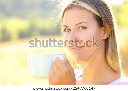 Satisfied woman looks at you holding coffee cup in a field