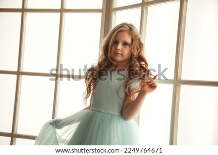 Portrait pensive little girl in art azure dress posing near large light window in room, looking at camera. Studio shot positive thoughtful kid lady. Child emotion concept. Copy text space for ad