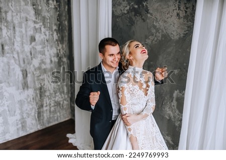 Stylish young groom in a blue suit and a beautiful blonde bride in a white lace dress are dancing, laughing while standing in the studio, interior on a gray wall background. wedding photography.