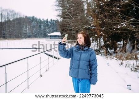woman in winter with a phone takes a selfie in nature in the park on a cold day
