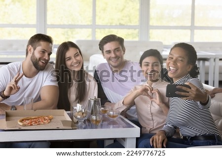 Happy multiethnic friends taking selfie on smartphone in cafe, shooting self video, holding mobile phone, smiling, laughing, posing at table with pizza, hot drink, having fun