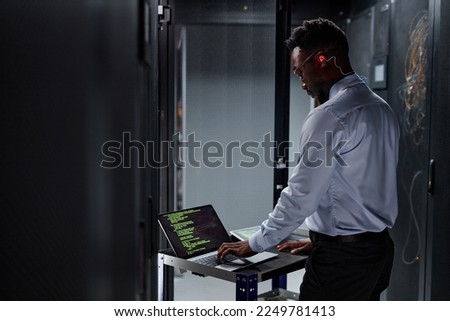 Side view portrait of black man as network engineer using laptop while setting up servers in data center Royalty-Free Stock Photo #2249781413