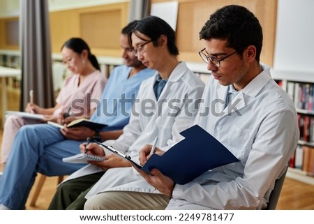 Group of medics in row taking notes during lecture or seminar, focus on young man holding clipboard in foreground Royalty-Free Stock Photo #2249781477