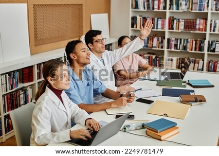 Diverse group of young med students sitting in row and raising hands during lecture or seminar Royalty-Free Stock Photo #2249781479