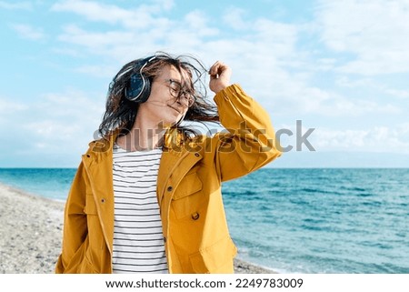 Smiling middle-aged female in headphones listening music and dancing while walking near the sea. Woman with closed eyes listening music or podcast from smartphone application in headphones in nature. Royalty-Free Stock Photo #2249783009