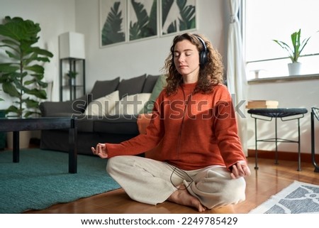 Young pretty healthy mindful woman wearing headphones listening calming music audio podcast sitting on floor at home doing yoga meditation relaxing exercise for mental balance feeling peace of mind. Royalty-Free Stock Photo #2249785429