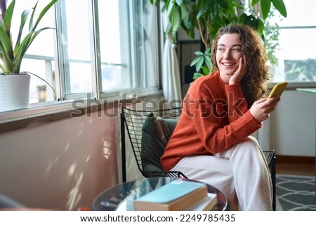 Happy young woman sitting on chair holding mobile phone using cellphone device, checking modern apps on smartphone, texting messages, browsing internet doing shopping relaxing at home. Royalty-Free Stock Photo #2249785435