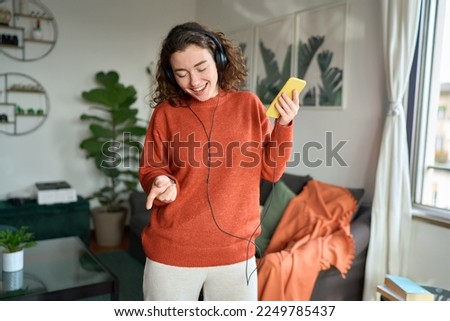 Happy pretty young woman wearing headphones using smartphone dancing at home. Cheerful girl listening music on mobile phone, singing song feeling relaxed standing in modern living room interior.