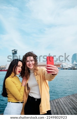 Two women taking selfies with a mobile phone outdoors on the sea promenade.