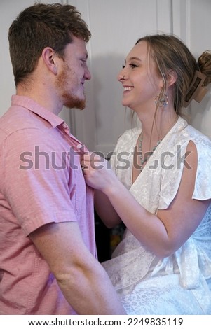 A young, caucasian 20-something couple poses for a portrait in their kitchen; the bride-to-be sits on the counter with the groom-to-be standing next to her Royalty-Free Stock Photo #2249835119
