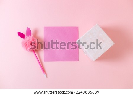 A gift box, a pink blank sheet, a pen with a fluffy head and bunny ears lie in the center on a pale pink background with copy space, flat close-up. Valentine's day concept. Royalty-Free Stock Photo #2249836689