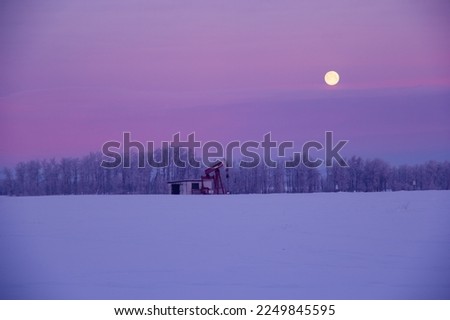 Winter landscape in prairies - field along the forest covered with snow and the pump jack in the center extracting oil, full moon in the sky