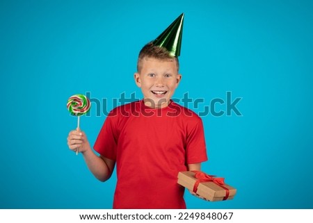 Portrait Of Joyful Birthday Boy Wearing Party Hat Holding Present And Hard Candy, Happy Preteen Male Child Celebrating His B-Day, Posing Isolated Over Blue Studio Background, Copy Space
