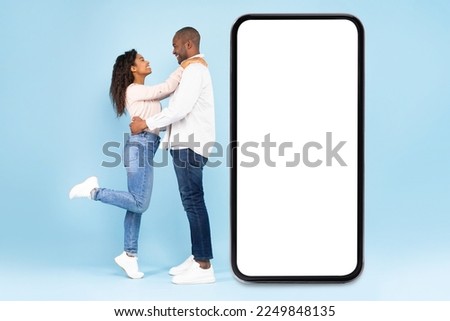 Loving black couple embracing and standing near large mobile phone with empty screen, advertising application, blue studio background. Full length, mockup