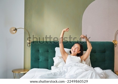 Portrait of smiling happy asian girl, wakes up feeling enthusiastic, stretches her hands up, enjoys good morning in her bedroom. Royalty-Free Stock Photo #2249860529
