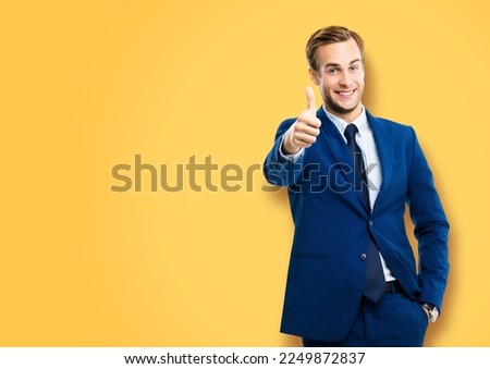 Image of excited businessman showing thumbs up like hand sign gesture, in blue suit, over vivid yellow colour background. Handsome happy man. Copy space for ad, slogan or text.