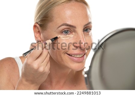 Middle aged senior woman applyes concealer under her eyes on white studio background Royalty-Free Stock Photo #2249881013